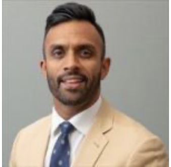 Man wearing beige suit jacket, white shirt, and blue tie, with short hair and beard, posing against a gray background.